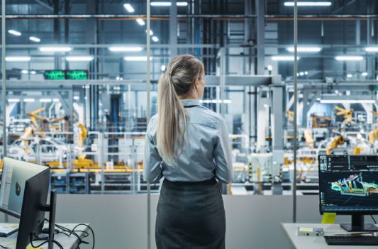 Car Factory Office: Successful Female Chief Engineer Overlooking Factory Production Conveyor. Automated Robot Arm Assembly Line Manufacturing Advanced High-Tech Electric Vehicles. Back View Shot